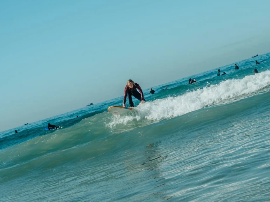 A surfer in a maroon wetsuit stands up on a white board as a blue wave breaks.